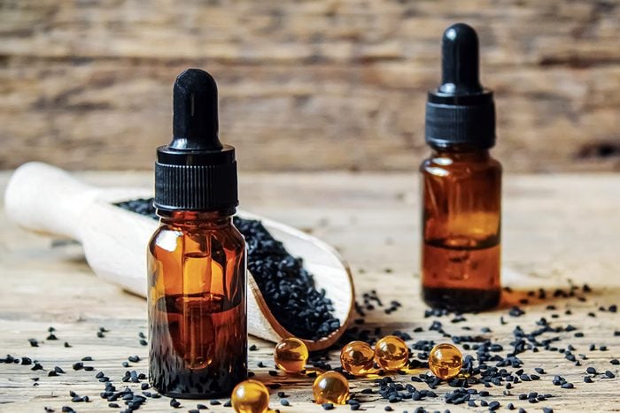 Two small amber dropper bottles sitting on a wooden surface next to a wooden scoop filled with black seeds and several yellow softgel capsules, with a rustic wooden background.