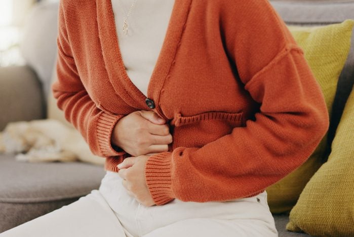 Stomach ache, cramps and hands of woman with abdomen pain due to constipation, menstruation or IBS issue. Sick, home and person suffering and holding belly in a house lounge, couch and living room