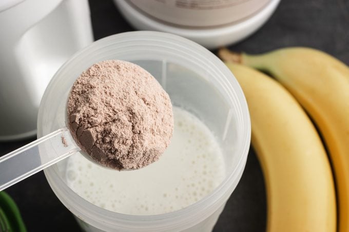 A plastic measuring spoon putting whey protein powder into a shaker with milk on a dark background with bananas, illustrating the process of making a healthy protein drink