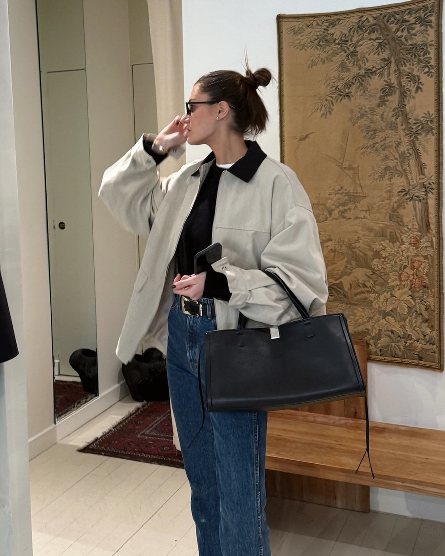 NYC style influencer Coco Schiffer poses in a store wearing a barn coat, simple black bag, black crewneck sweater layered over a white t-shirt, a belt, and jeans