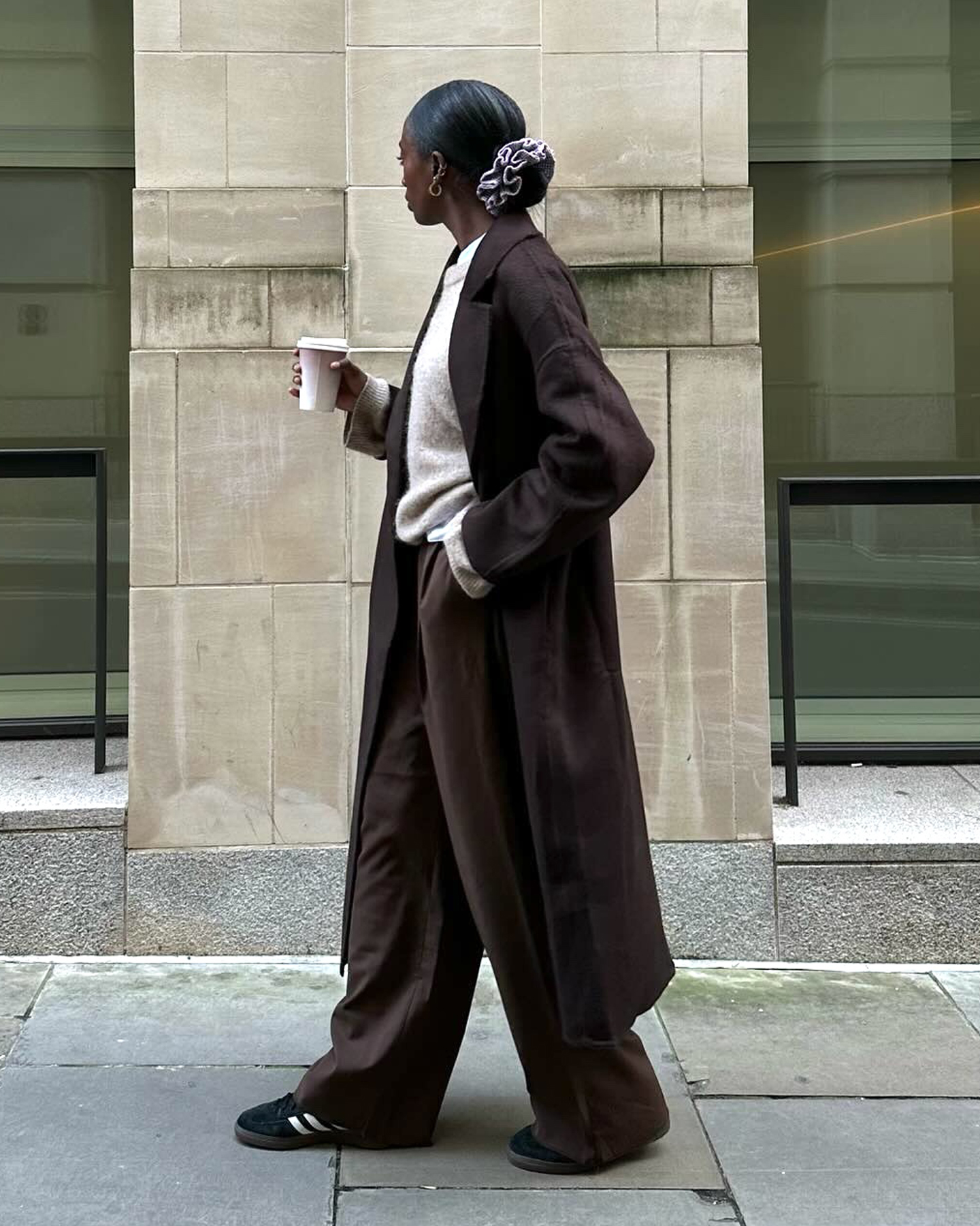 London style influencer Liv Madeline poses with a ruffled scrunchie, long brown coat, a neutral sweater, brown relaxed pants and black Adidas sneakers