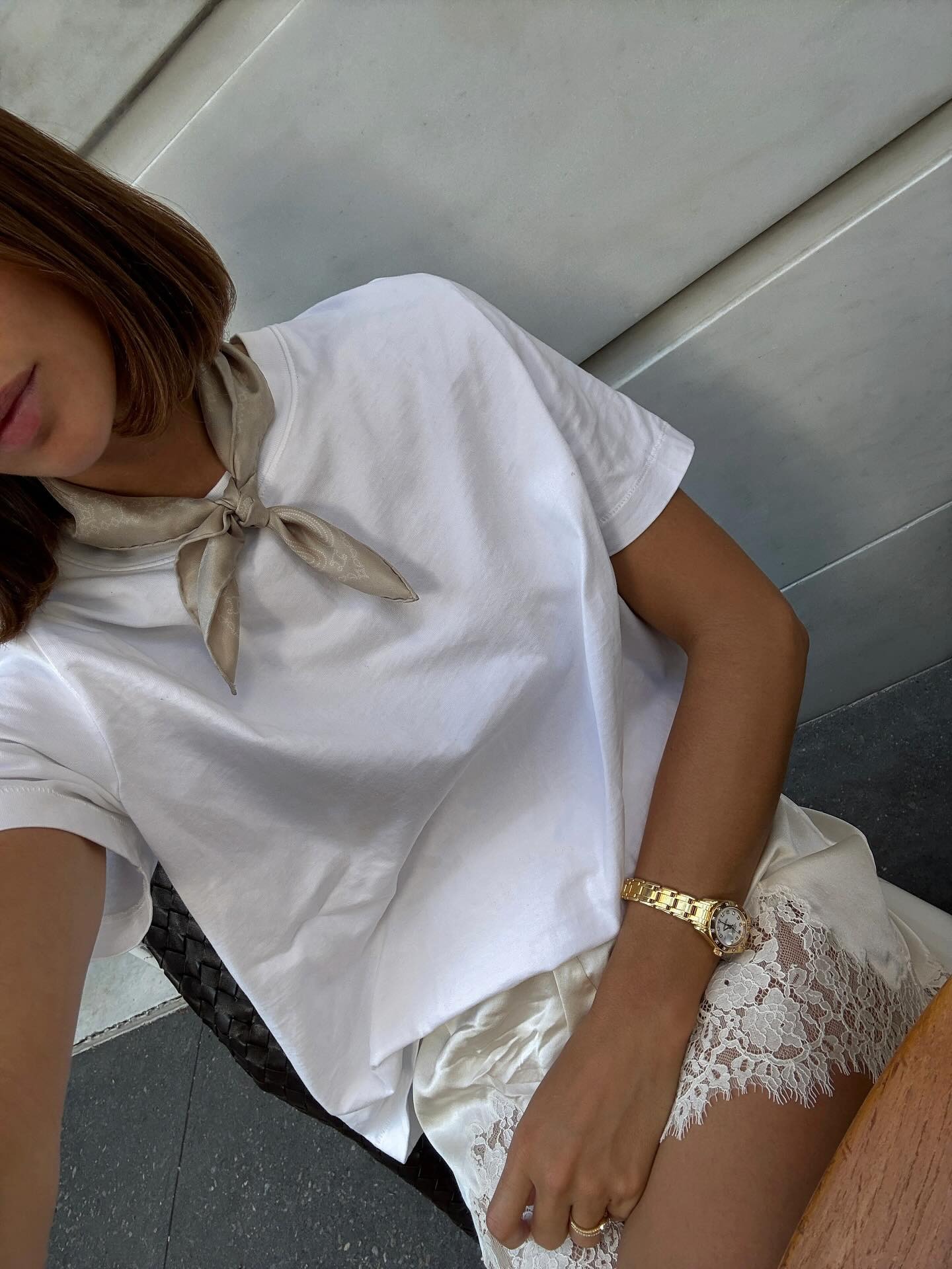 Woman takes a selfie in a white t-shirt, white lace satin shorts, beige neck scarf, and gold watch.