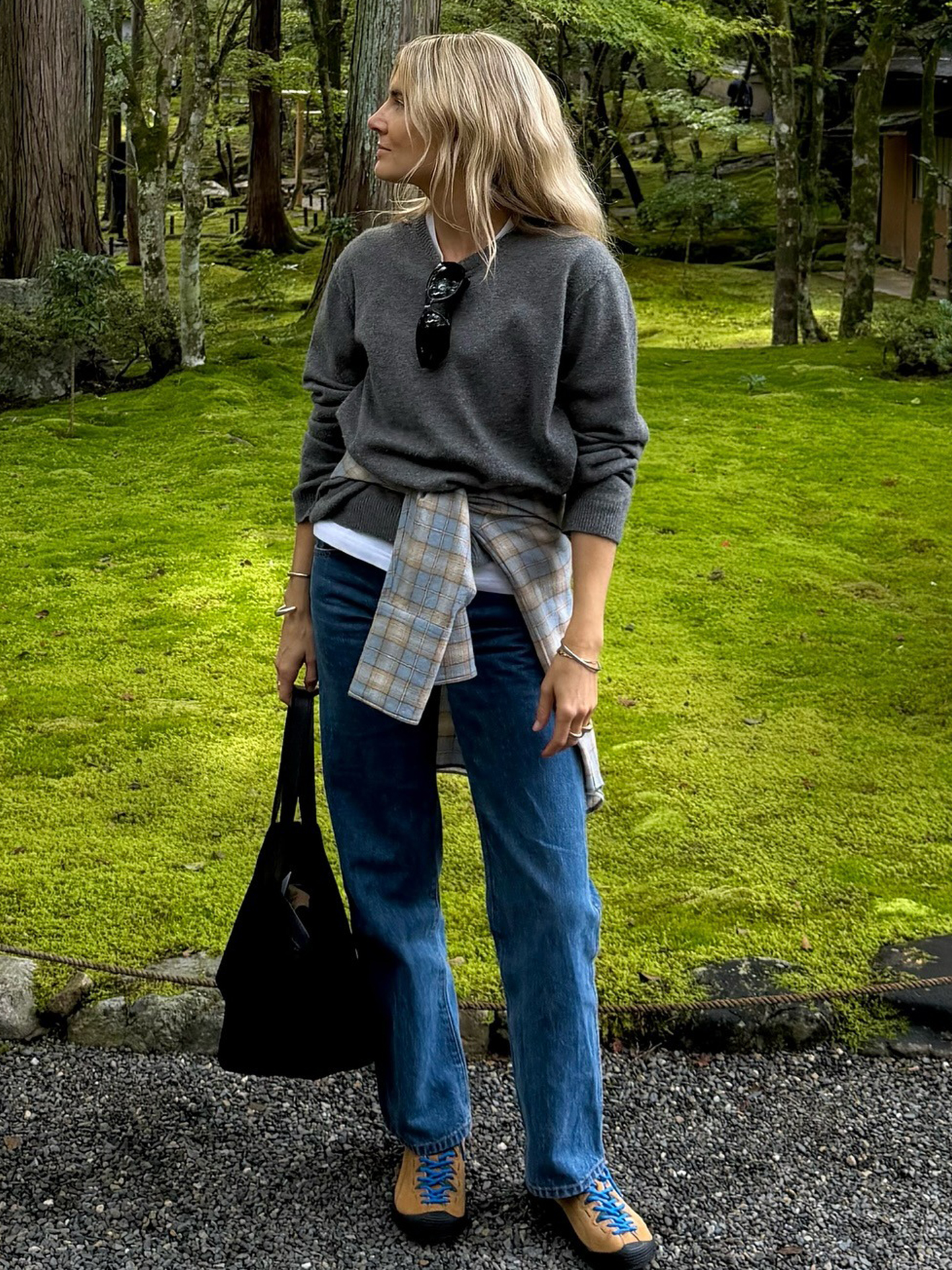 British Lucy Williams visiting Japan wearing a gray sweater layered over a white T-shirt, a plaid shirt wrapped around her waist, straight-leg jeans, and Keen Jasper sneakers with blue laces.