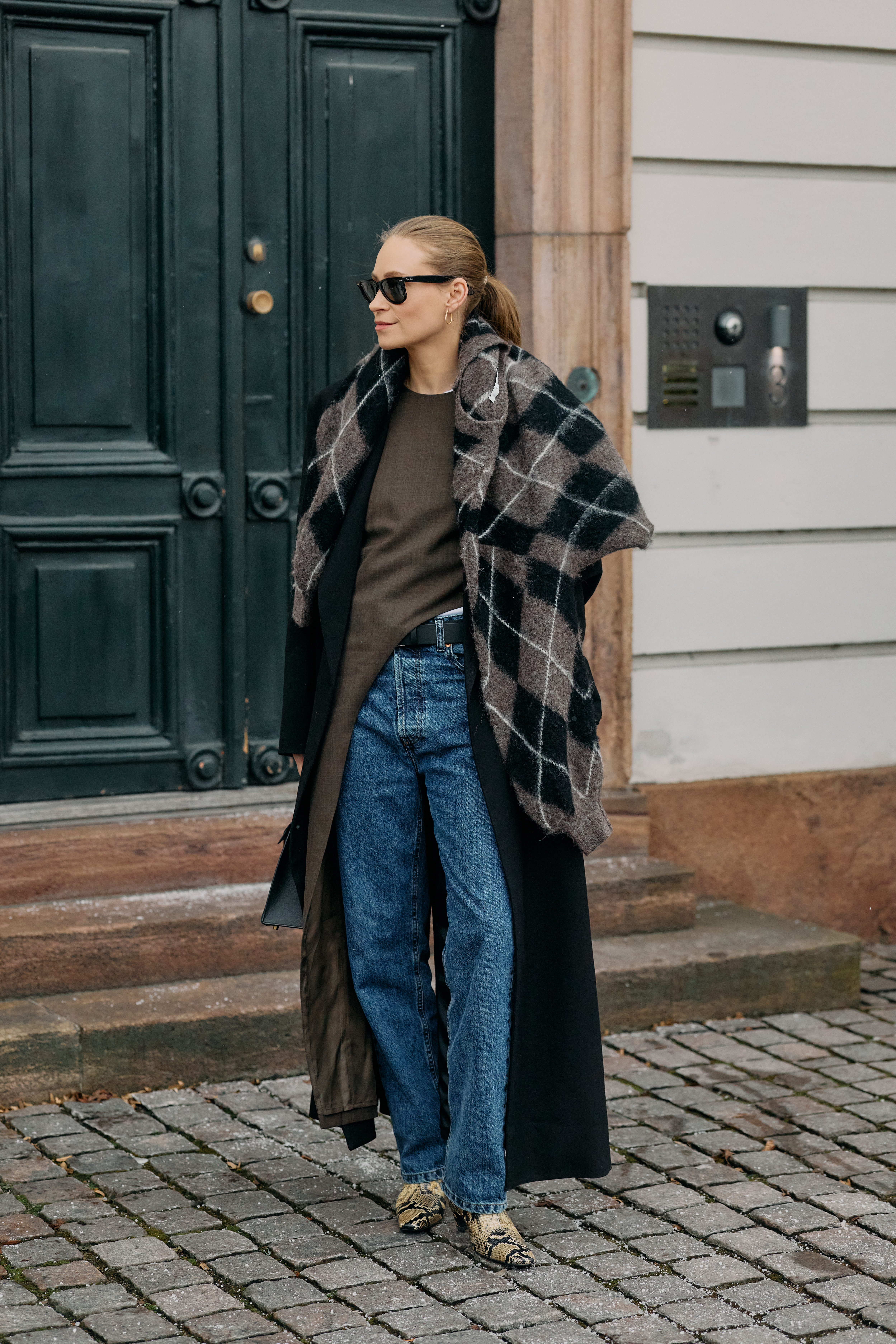 A woman wears a plaid scarf, stovepipe jeans, and snakeskin shoes.