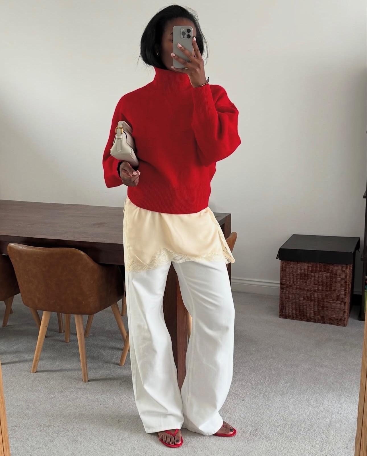 British style influencer Marilyn Nwawulor-Kazemaks poses in her London dining room with a red turtleneck sweater layered over an ivory lace-trim top, white jeans, and red flip-flop sandals