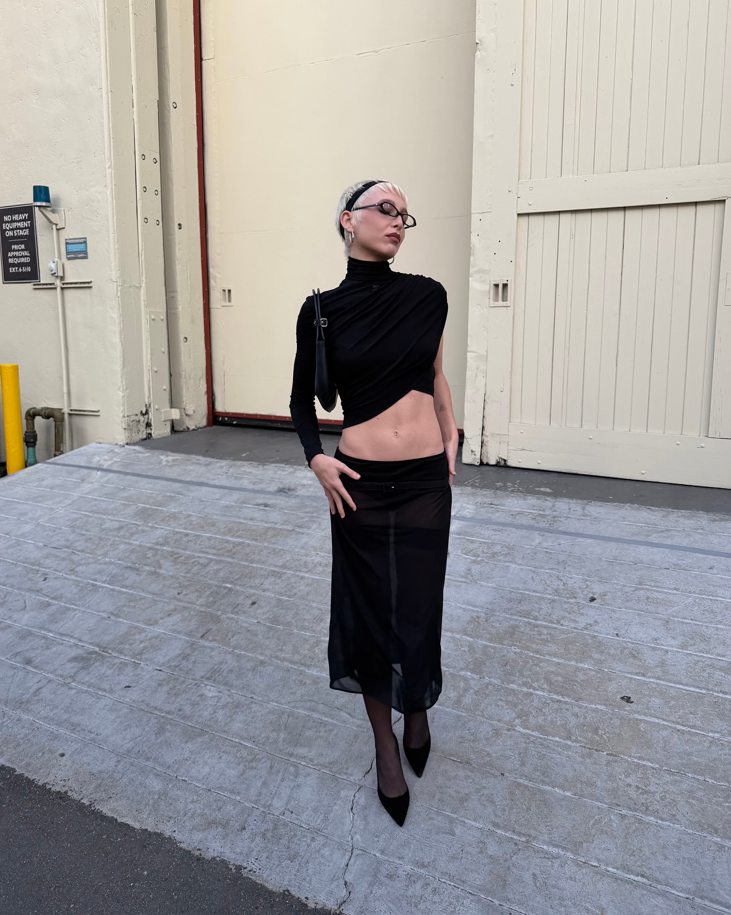 Emma Chamberlain is wearing a sheer black skirt, tights, a black long sleeve, and pointed-toe heels