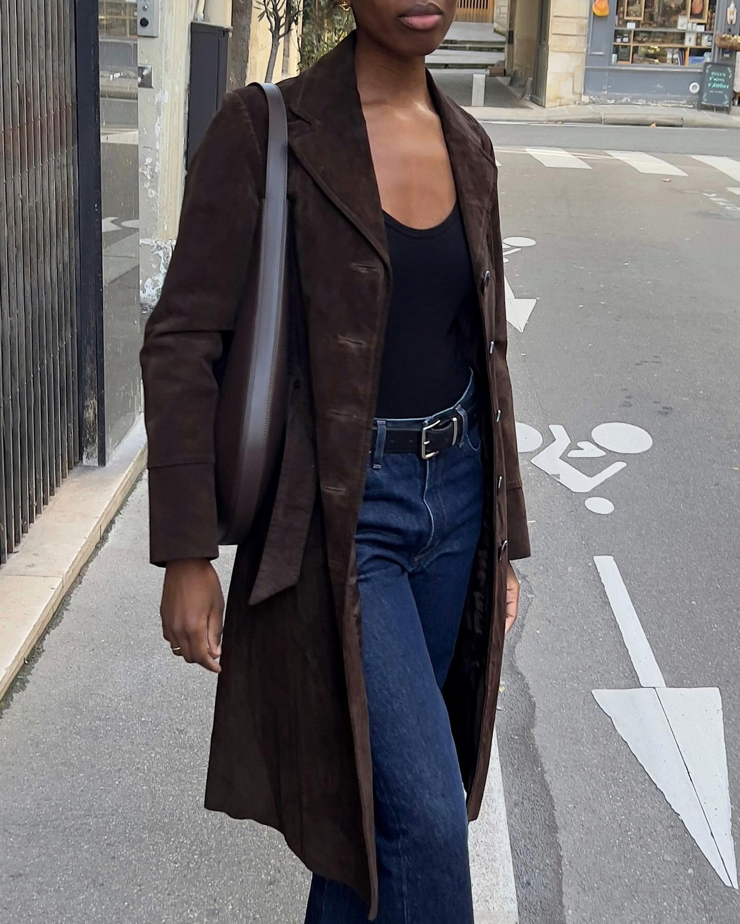 European style influencer Sylvie Mus poses in a brown suede coat, a black top, and dark-wash jeans